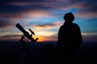 Bir adamın silueti, teleskop ve kırsal alan yıldızlı gökyüzünün altında.