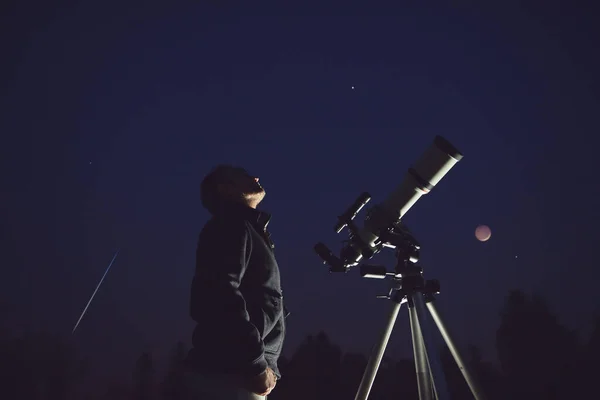 Silhouette of a man, telescope, stars, planets and shooting star under the night sky.