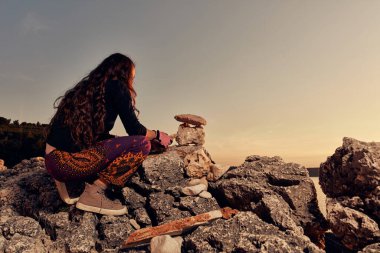 Gün batımında okyanus kıyısında kaya ve taşları dengeleyen bir kadının silueti..