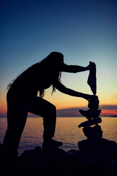 Silhouette of a woman balancing rocks and stones on the ocean sea coast at sunset sunrise time.