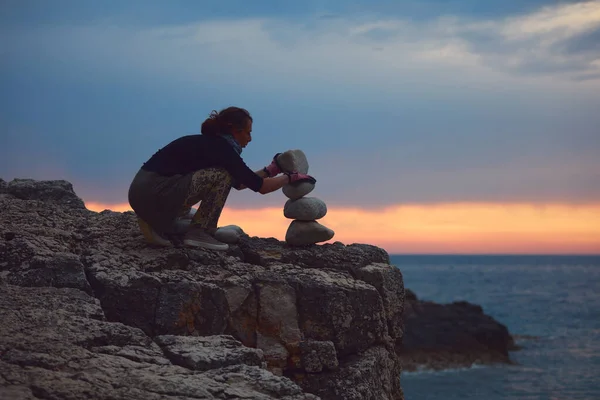 Silhouette of a woman balancing rocks and stones on the ocean sea coast at sunset sunrise time.