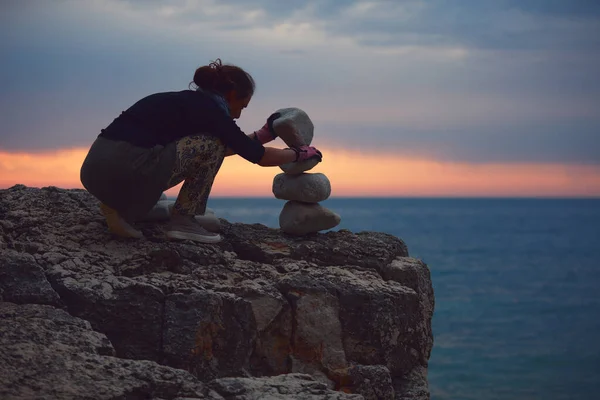 Silhouette of a woman balancing rocks and stones on the ocean sea coast at sunset sunrise time.