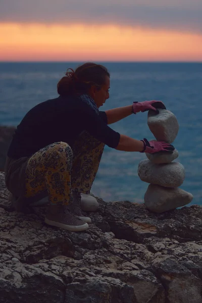 Silhouette of a woman balancing rocks and stones on the ocean sea coast at sunset sunrise time.