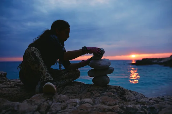 Silhouette of a woman balancing rocks and stones on the ocean sea coast at sunset sunrise time.