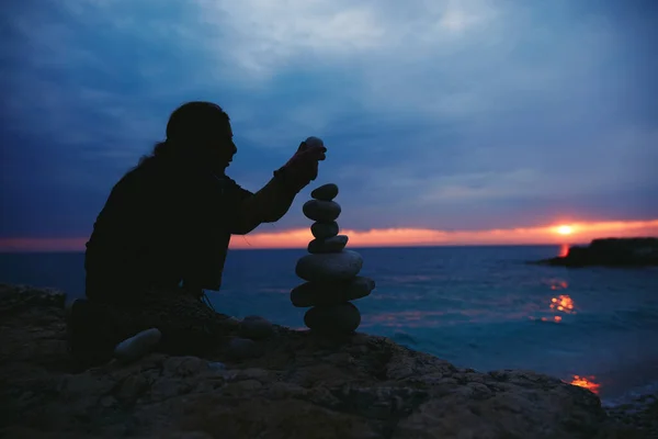 Silhouette of a woman balancing rocks and stones on the ocean sea coast at sunset sunrise time.