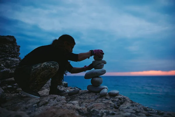 Silhouette of a woman balancing rocks and stones on the ocean sea coast at sunset sunrise time.