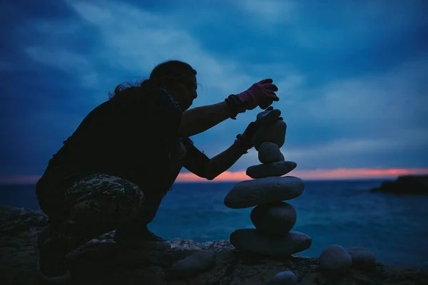 Silhouette of a woman balancing rocks and stones on the ocean sea coast at sunset sunrise time.