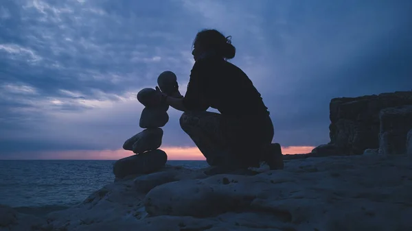 Silhouette of a woman balancing rocks and stones on the ocean sea coast at sunset sunrise time.