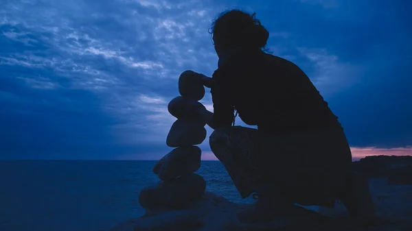 Silhouette of a woman balancing rocks and stones on the ocean sea coast at sunset sunrise time.