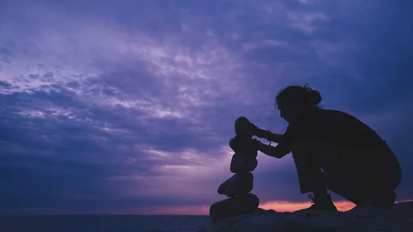 Silhouette of a woman balancing rocks and stones on the ocean sea coast at sunset sunrise time.