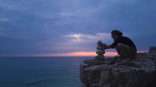 Silhouette of a woman balancing rocks and stones on the ocean sea coast at sunset sunrise time.