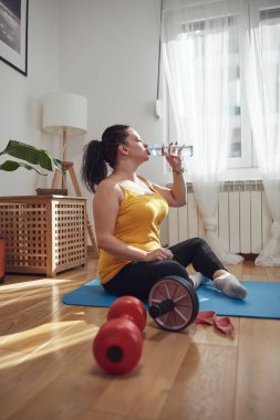 Kadın oturma odasında antrenman yapıyor, evde egzersiz yapıyor, su içiyor ve duruyor..
