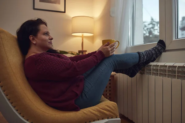 Woman heating feet on a chilly winter day, holding coffee tea cup, energy and gas crisis, cold room, heating problems.