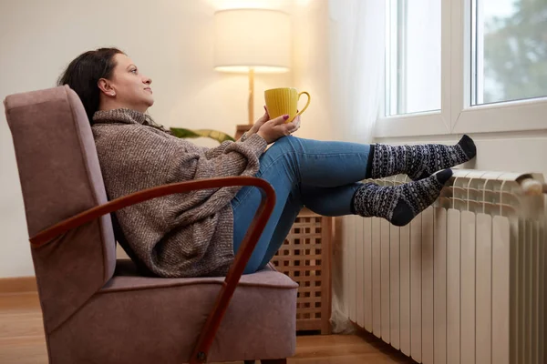 Woman heating feet on a chilly winter day, holding coffee tea cup, energy and gas crisis, cold room, heating problems.