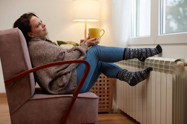 Woman heating feet on a chilly winter day, holding coffee tea cup, energy and gas crisis, cold room, heating problems.