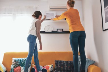 Anne ve kızı kanepede duruyor, resimlerini ve fotoğraflarını evdeki raflara asıyorlar..