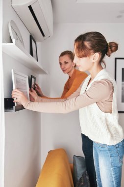 Anne ve kızı kanepede duruyor, resimlerini ve fotoğraflarını evdeki raflara asıyorlar..