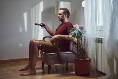 Yetişkin bir adam evde televizyon izleyip uzaktan kumanda tutuyor..