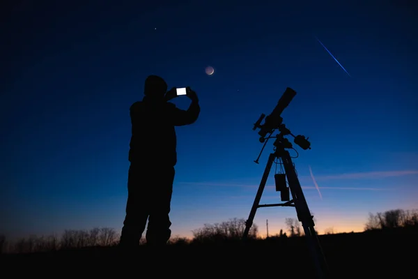 Silhouette of a man with smartphone, telescope and countryside under evening skies.