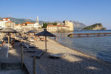 Karadağ - Budva: 9 Eylül 2021: Plaza Ricardova Glava plajı ve dağ manzaralı güneş yataklarında yatan çok sayıda insan. Budva 'nın ünlü plajı, eski kasaba ve Balkan dağları manzarası.