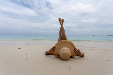 Hasır şapka takan genç bir kadın yaz tatili konsepti olan Phuket, Tayland 'da kumsalda bacaklarını uzatıyordu..