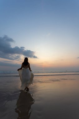 Beyaz elbiseli mutlu kadın sahilde gün batımının tadını çıkarıyor.