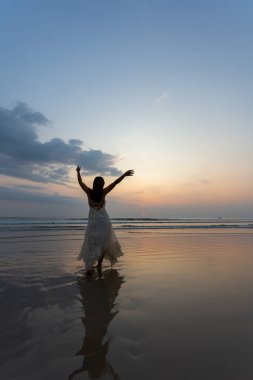 Beyaz elbiseli mutlu kadın sahilde gün batımının tadını çıkarıyor.