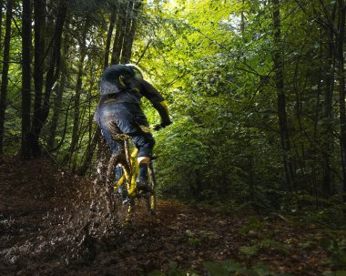 Cyclist in full face helmet on the yellow enduro bicycle fast rides in the mud in a green forest. Summer time, Ukraine
