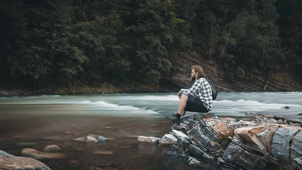 A man with a beard in a plaid shirt and shorts sits on the bank of a mountain river, moody colors Ukraine