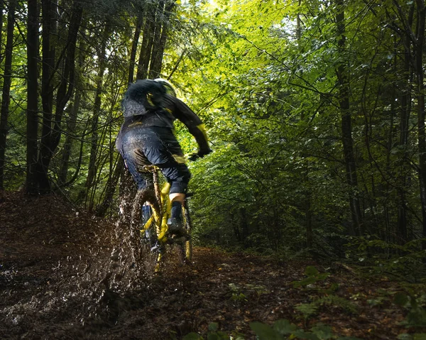 Cyclist in full face helmet on the yellow enduro bicycle fast rides in the mud in a green forest. Summer time, Ukraine