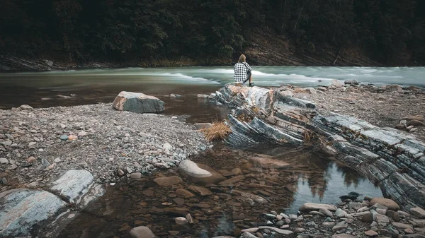 A man with a beard in a plaid shirt and shorts sits on the bank of a mountain river, moody colors Ukraine