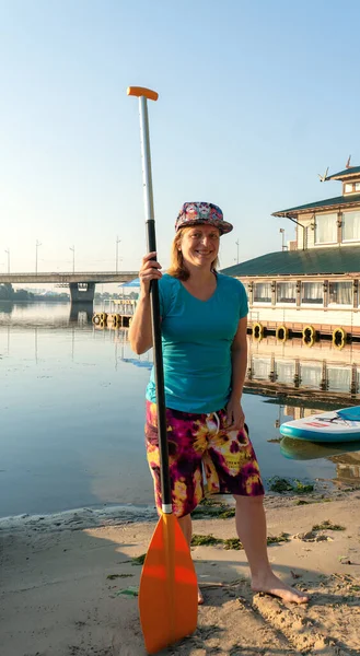 A young girl wearing a full cap, blue T-shirt and board short stands with a paddle, Kyiv, Ukraine