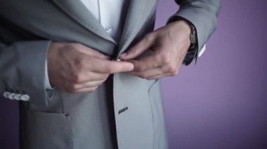 Man's hands fasten buttons on grey classic jacket against purple wall. A close-up of a young man's hands in a classic grey jacket, white shirt and watch on his hand.