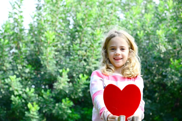 Beautiful blond girl sittig o a tree branch holding large felt heart in her hands — Stock Photo, Image