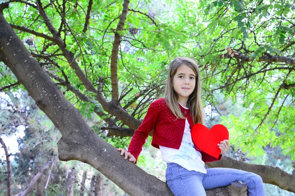 Hermosa chica rubia sentada en una rama de árbol sosteniendo un gran corazón de fieltro en sus manos — Foto de Stock
