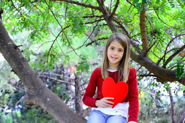 Hermosa chica rubia sentada en una rama de árbol sosteniendo un gran corazón de fieltro en sus manos —  Fotos de Stock