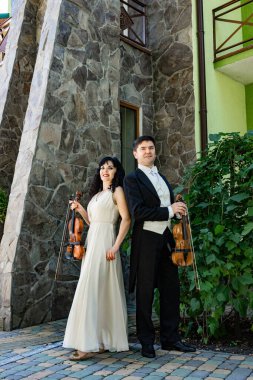 male and female violinists stand in concert costume with paper clips in their hands near the old house