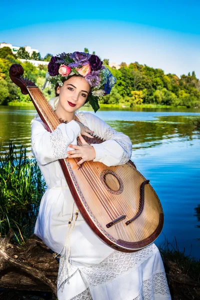 Genç bir Ukraynalı kadın güneşli bir günde nehir kenarında oturur ve banduraya sarılır..