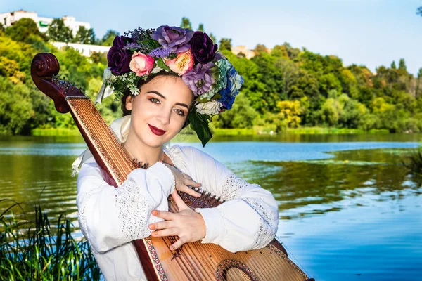 A young Ukrainian woman sits by the river on a bright sunny day and hugs the bandura — Stock fotografie