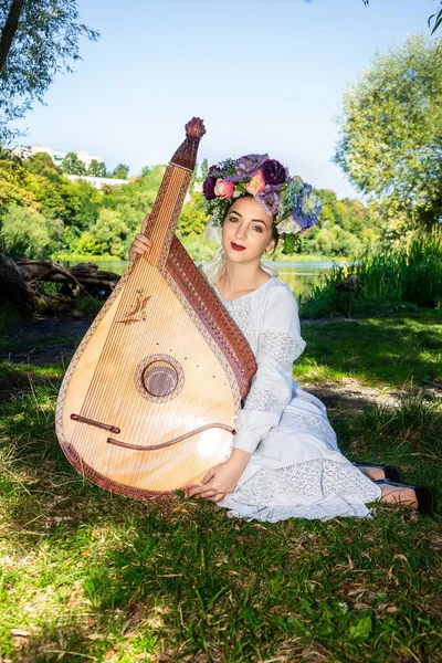 Ukraynalı genç bir kadın ulusal elbisesiyle nehir kenarında oturuyor ve elinde Ukrayna ulusal bandura enstrümanı tutuyor.