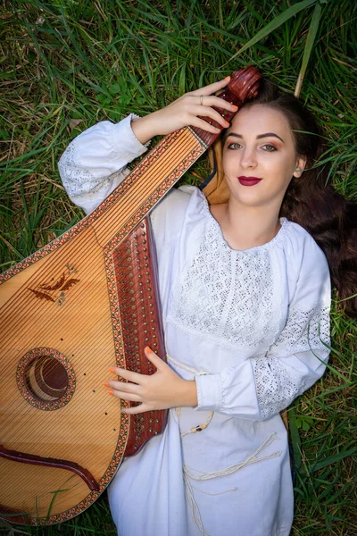 身穿白色绣花裙的乌克兰女孩躺在草地上，用乌克兰国家管弦乐队乐器演奏 — 图库照片