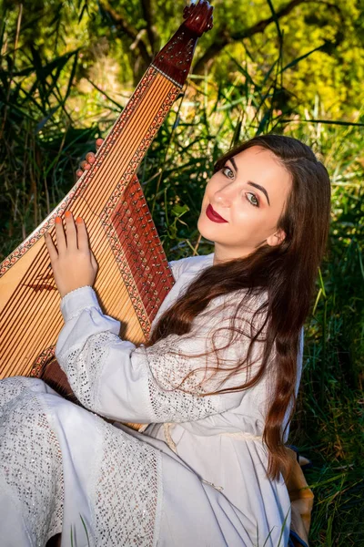 Joven mujer ucraniana en un vestido blanco y con el pelo que fluye se sienta en la hierba verde y sostiene una bandura — Foto de Stock