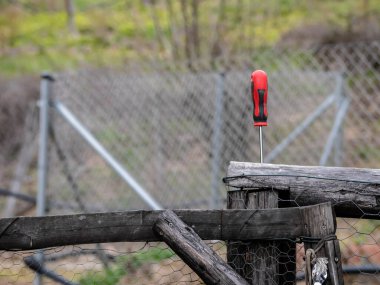 Kırmızı saplı tornavida bozulmuş ahşap bir çite çivilenmiş ve arka planda bir meyve bahçesi ve bitki örtüsünü koruyan metal ağlarla korunmuş.