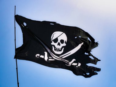 Tamamen parçalanmış ve morarmış siyah korsan bayrağı (Jolly Roger) rüzgarda dalgalanan bir bayrağa bağlanmış, kafatası yaması ve kumaşın üzerine beyaz çizilmiş iki kılıçla birlikte.