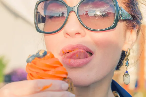 A woman in sunglasses licks her lips in orange cupcake cream.