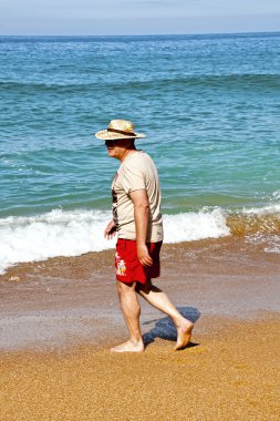 Deniz kenarında bir hasır şapka giyen adam yürüyor.