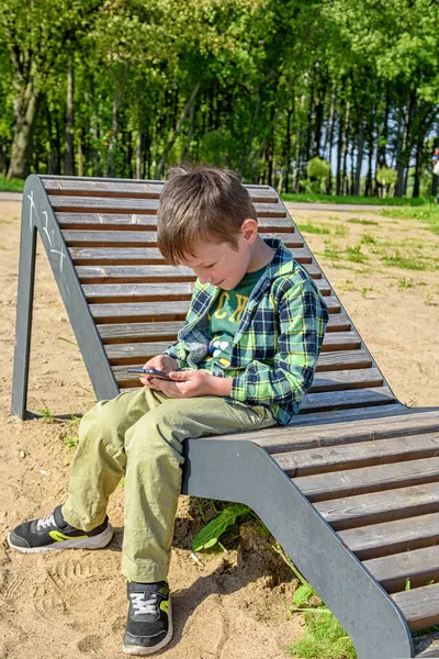 Feliz Niño Está Sentado Banco Jugando Teléfono Móvil Fondo Parque — Foto de Stock