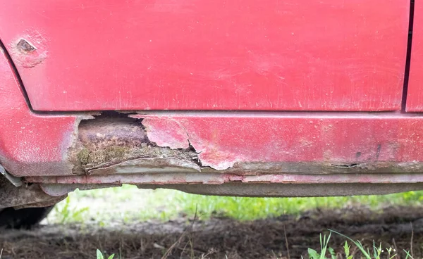 Rusty driver's door sills. Corrosion of the body of a red old car after winter. Influence of reagents in winter on an unprotected vehicle body. Damage to the left side, rotten threshold on the bottom