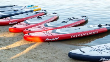 Boş SUP sörf tahtaları sakin bir nehirde ya da gölde sallanıyor. Su sporları. Kiralık dükkanda bir sürü tahta var. Suyun dalgalarından yansıyan güneş. Çeviriyorum. Ukrayna, Kyiv - 07 Ağustos 2022