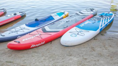 Boş SUP sörf tahtaları sakin bir nehirde ya da gölde sallanıyor. Su sporları. Kiralık dükkanda bir sürü tahta var. Suyun dalgalarından yansıyan güneş. Çeviriyorum. Ukrayna, Kyiv - 07 Ağustos 2022